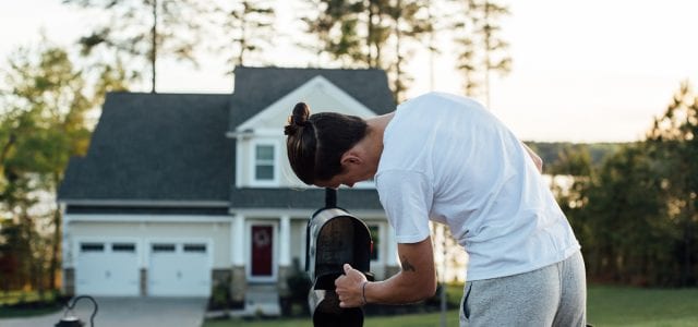 A young man in a white t-shirt checks his mailbox for letters and postcards in front of his house.