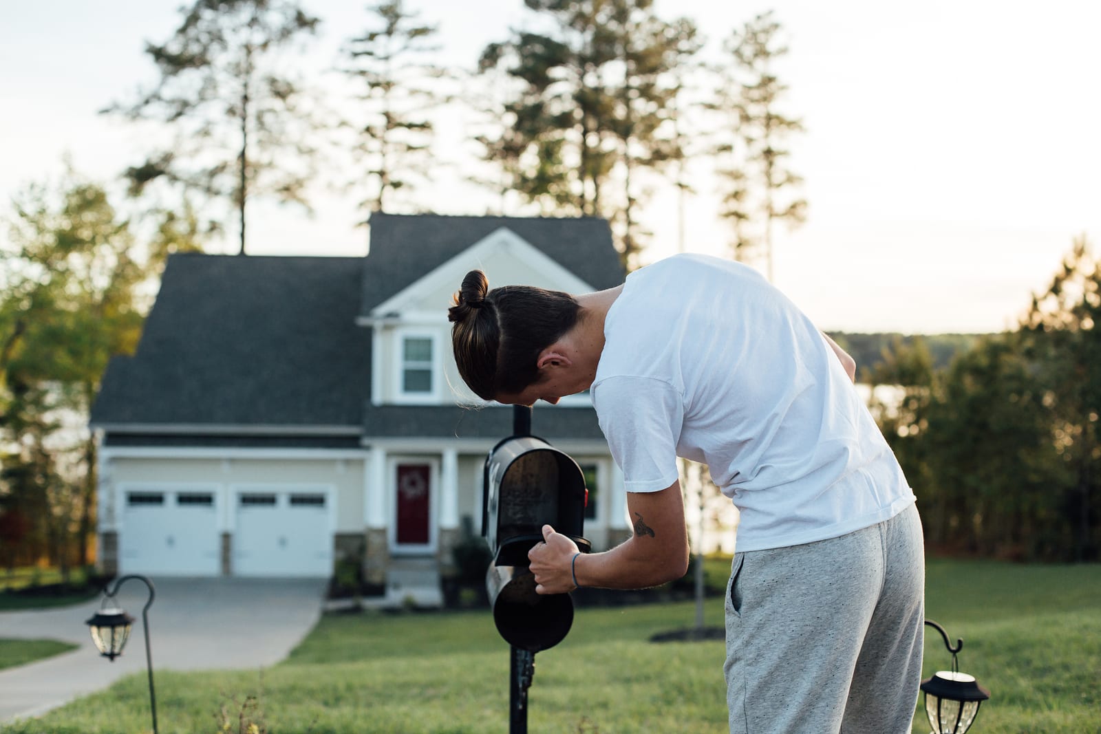 A young man in a white t-shirt checks his mailbox for letters and postcards in front of his house.