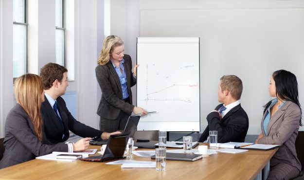 Blonde female present graph on flipchart during business meeting Blonde female present graph on flipchart during business meeting, while 4 more colleagues sits at conference table.