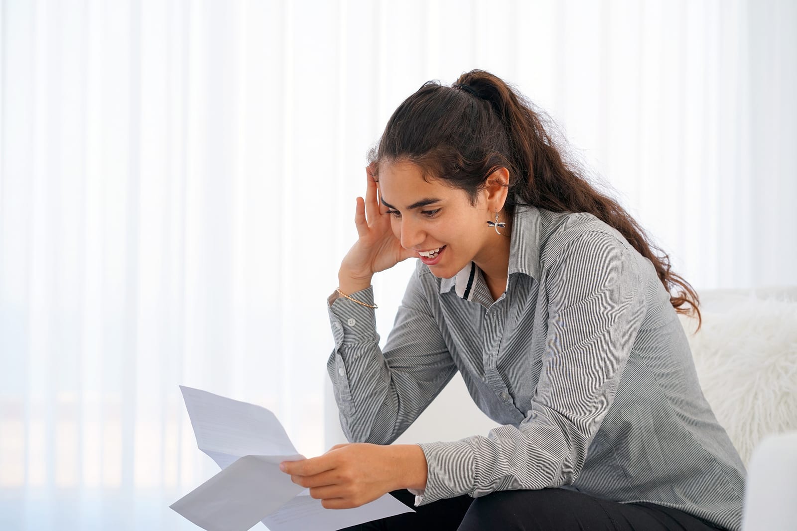 Happy young woman enjoying good news in writing. The girl reads a letter with good news sitting on the couch. An euphoric girl is happy after reading good news in a written letter, approving a loan.