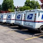 A line of USPS trucks are parked in a parking lot waiting for deliveries.