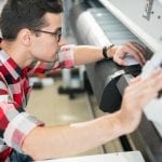 Serious thoughtful handsome young engineer in shirt examining wide format printer while holding inspection in printing house